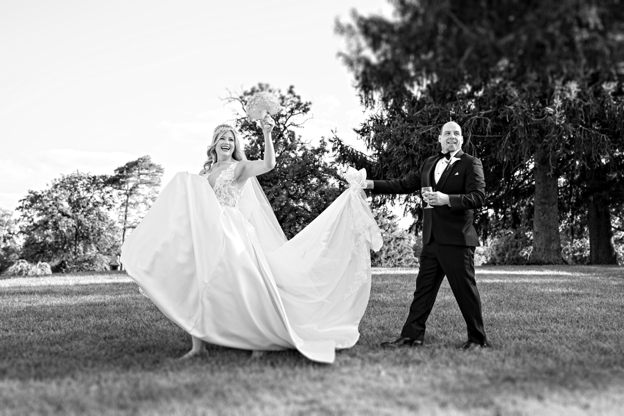 A groom helping with his brides wedding dress as they wave hello to their wedding guests.